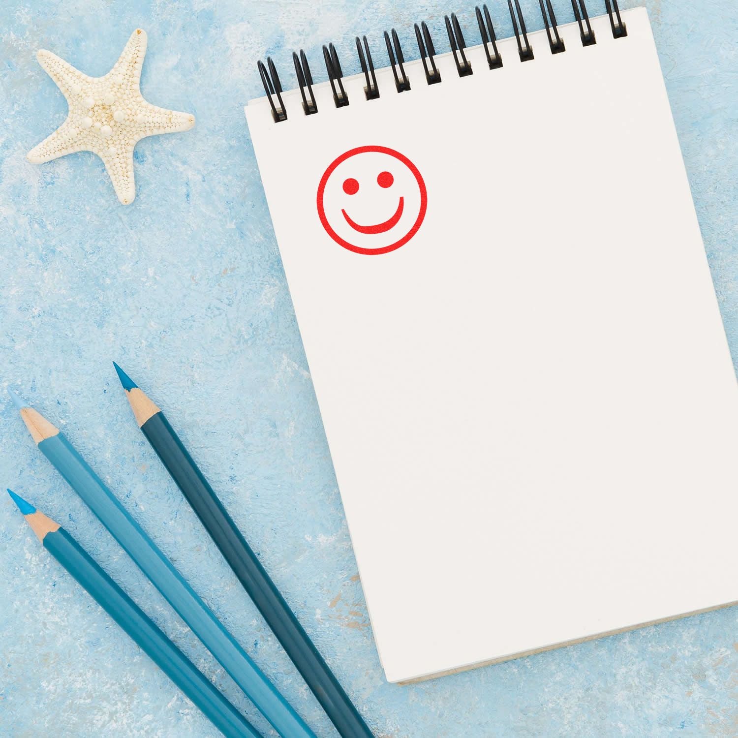 Round Smiley Rubber Stamp in red ink on a blank notepad, with blue pencils and a starfish on a light blue background.