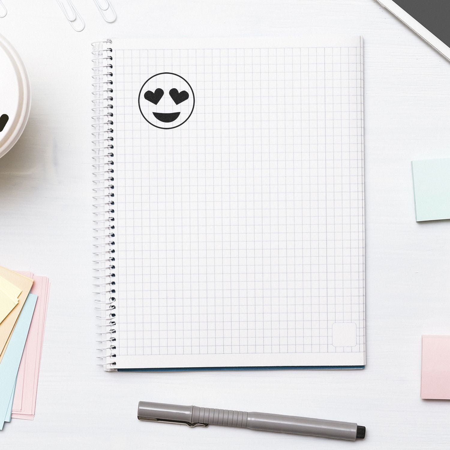 Round Smiley with Hearts Rubber Stamp on a grid notebook page, surrounded by a pen, sticky notes, and a smartphone.