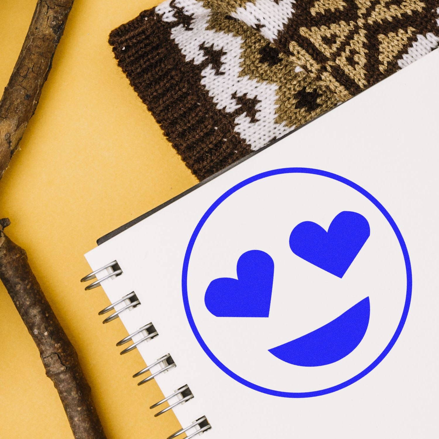 Round Smiley with Hearts Rubber Stamp on a notebook, next to a patterned knit sleeve and wooden sticks on a yellow background.