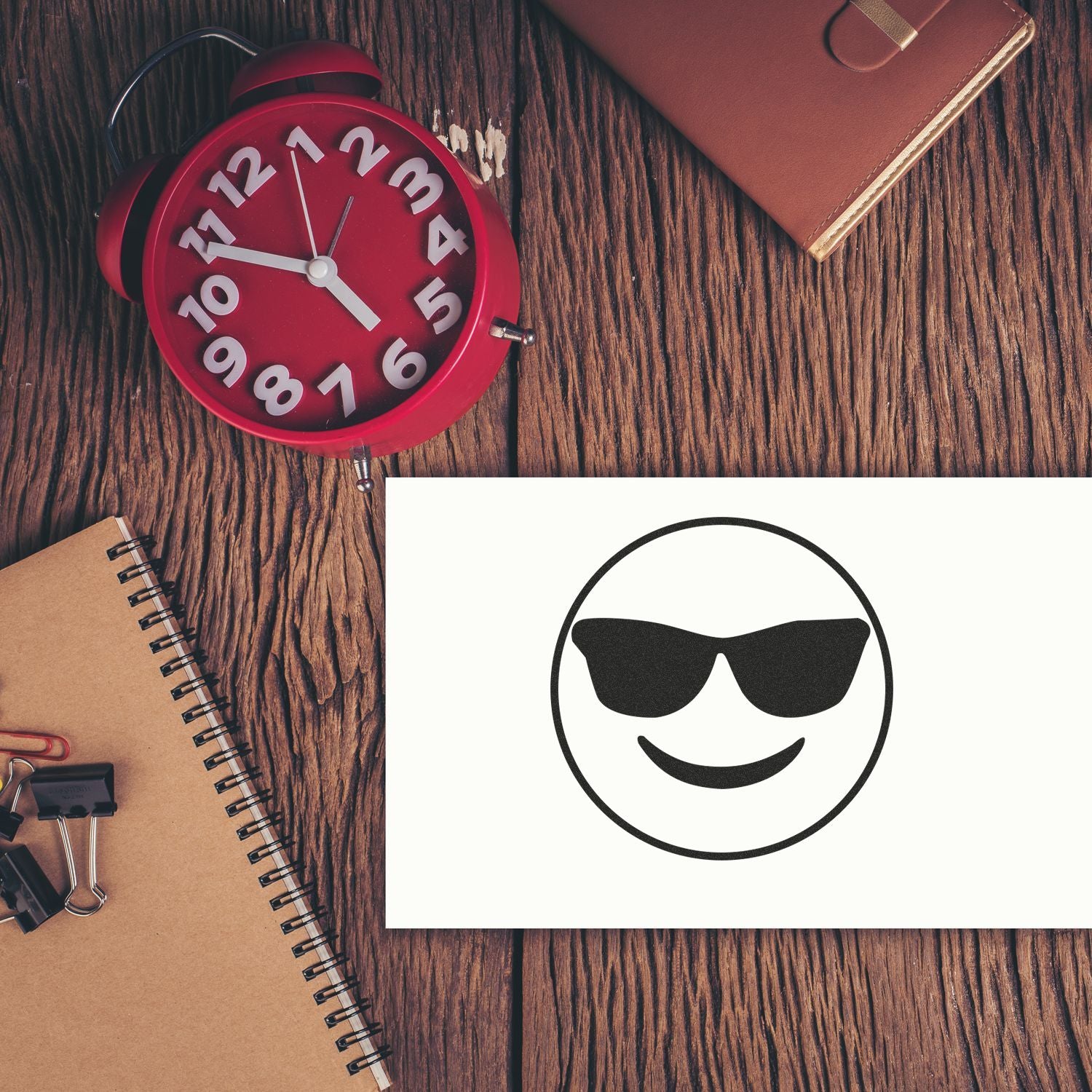 Round Smiley with Sunglasses Rubber Stamp on paper, placed on a wooden desk with a red clock, notebook, and office supplies.