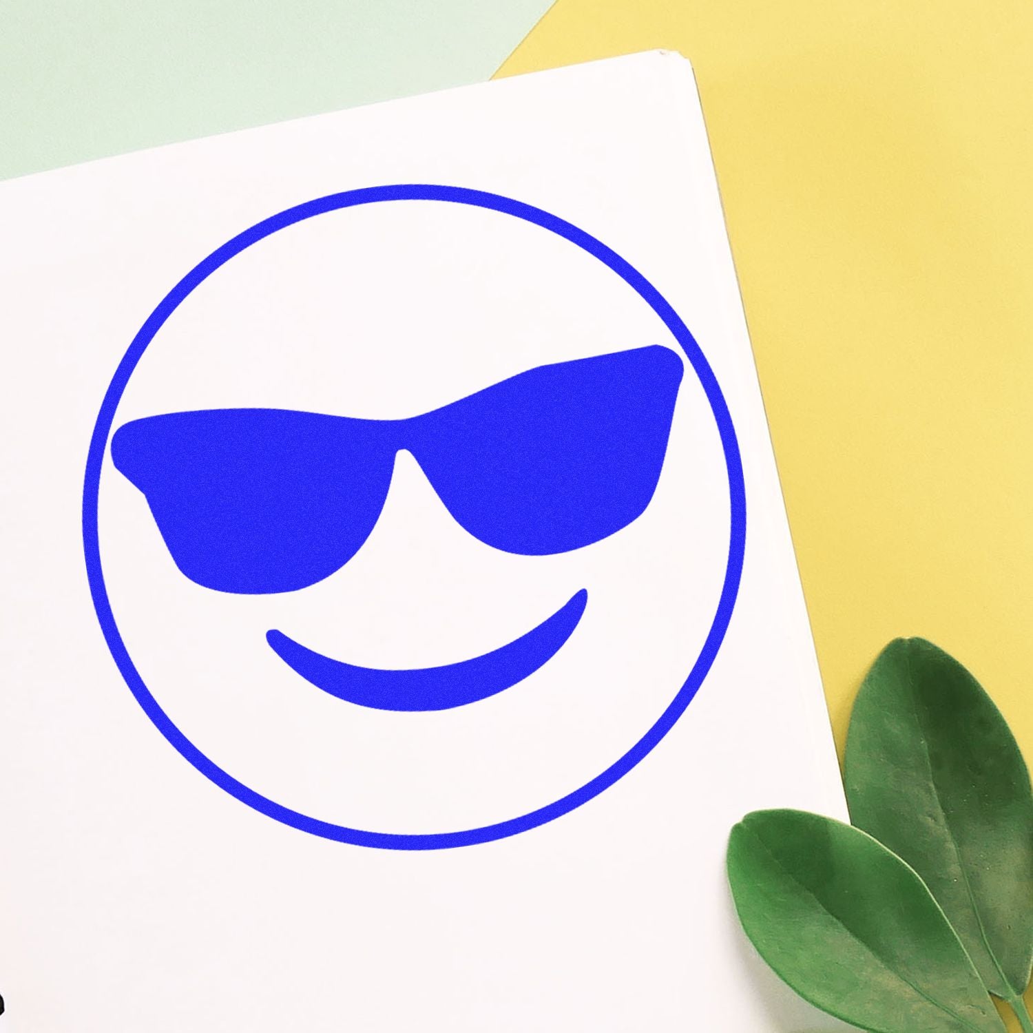 Round Smiley with Sunglasses Rubber Stamp in blue ink on white paper, with green leaves and yellow background.