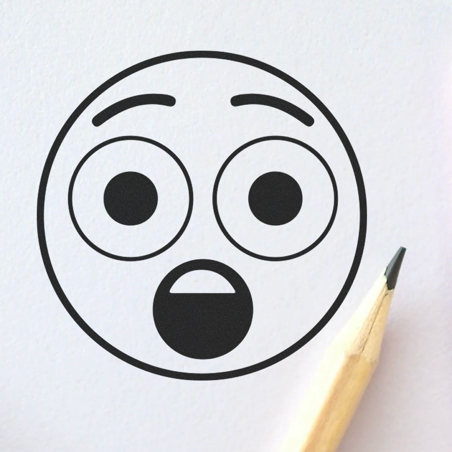 Round Surprised Smiley Rubber Stamp impression on white paper with a pencil tip pointing towards it.