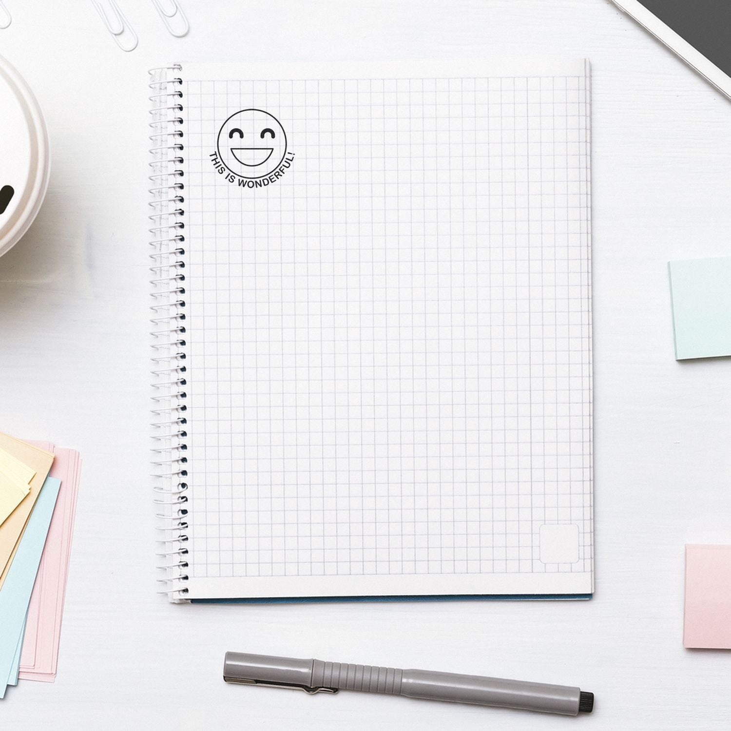 Round This is Wonderful Smiley Rubber Stamp on a spiral notebook with grid paper, surrounded by sticky notes and a pen.