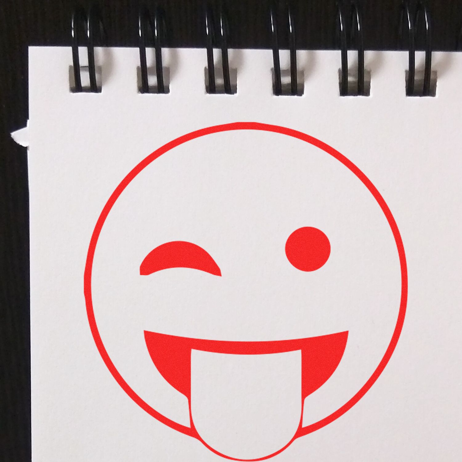 Round Tongue Out Smiley Rubber Stamp impression on a white notepad with black spiral binding.