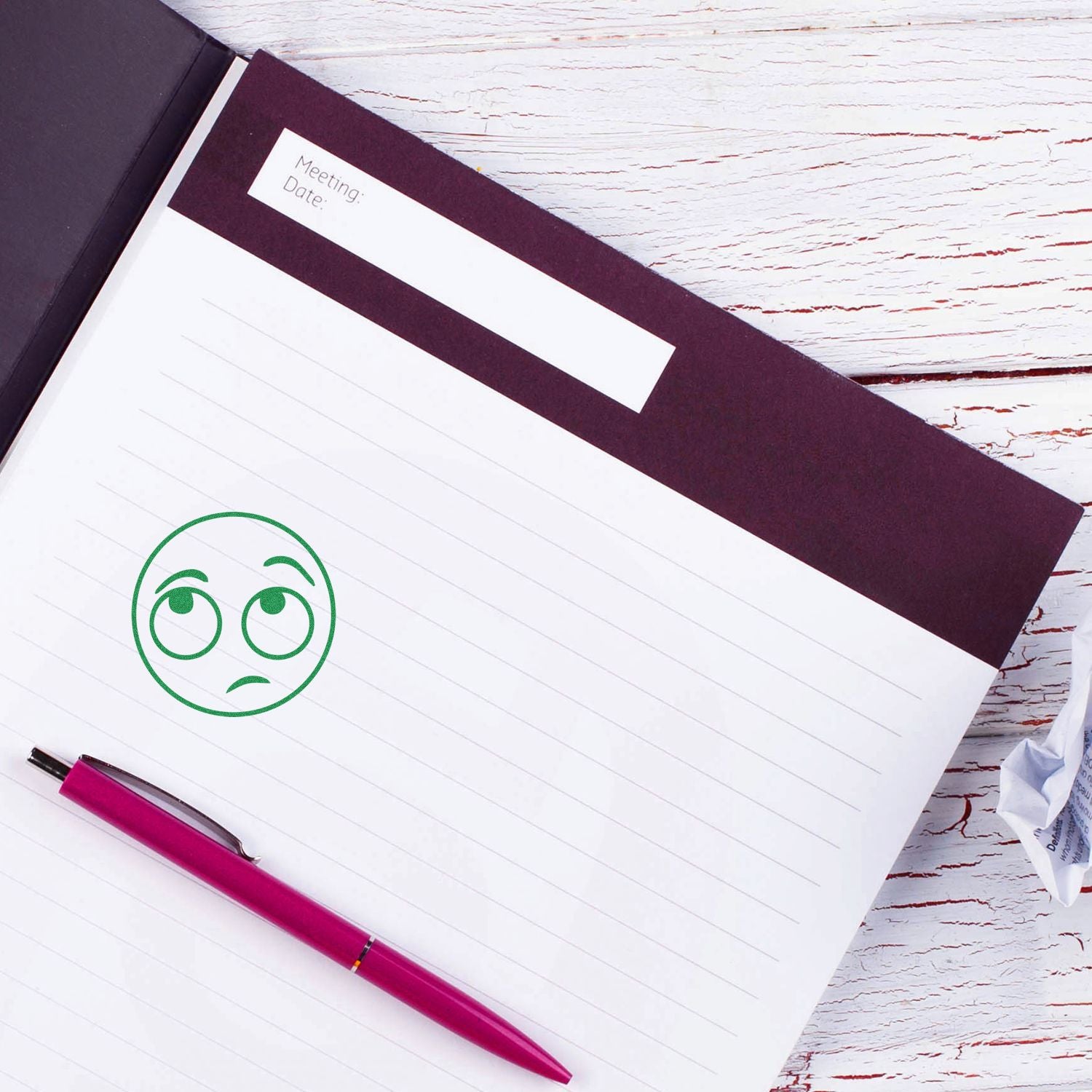 Notebook with a green Round Whatever Smiley Rubber Stamp impression, a pink pen, and a crumpled paper on a white wooden table.