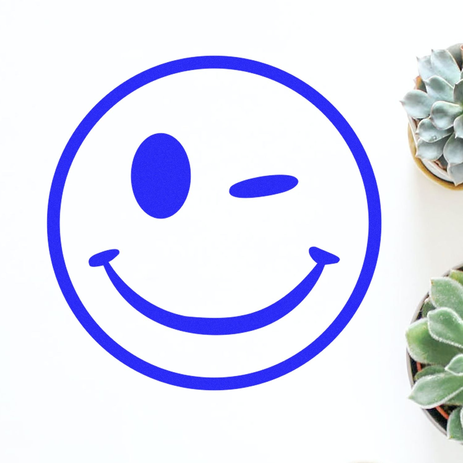 Round Winking Smiley Rubber Stamp in blue ink on white paper, surrounded by small potted succulents.