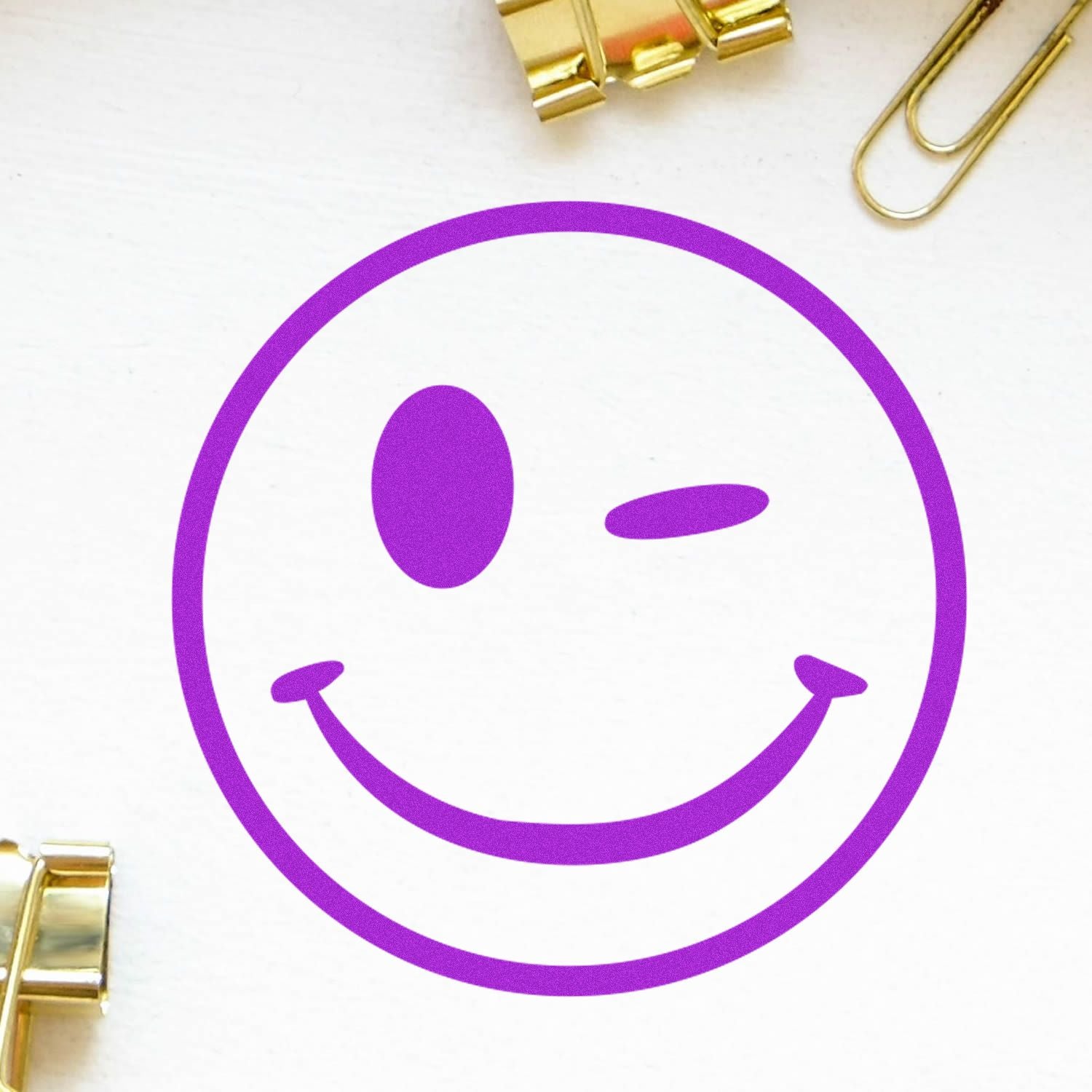 Round Winking Smiley Rubber Stamp in purple ink on white paper, surrounded by gold paper clips and binder clips.