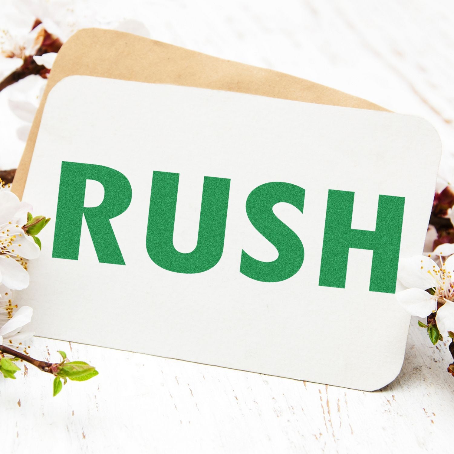 Self Inking Rush Stamp in use, stamping RUSH in bold green letters on a white card, surrounded by white flowers and a brown envelope.