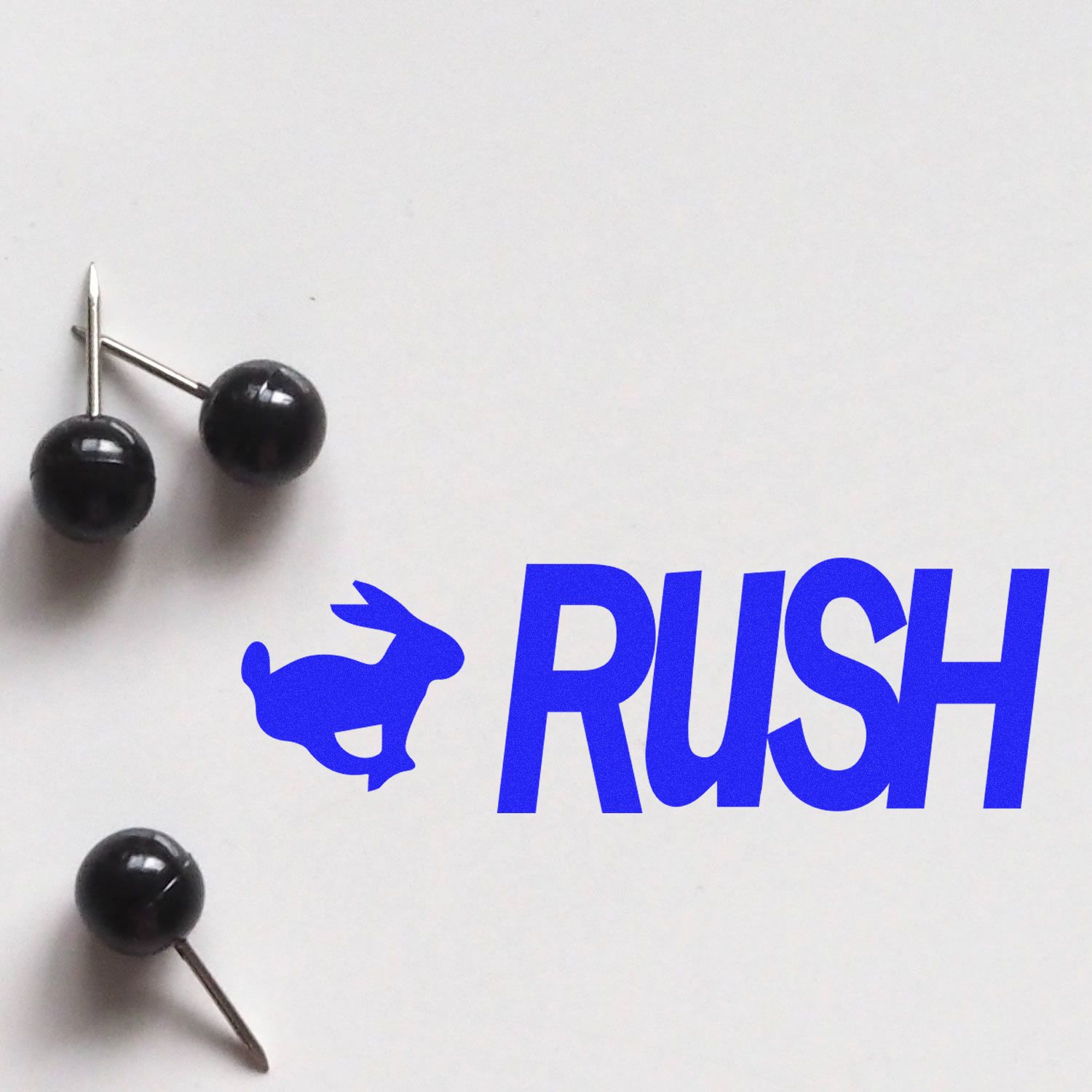 Rush with Rabbit Rubber Stamp in blue ink next to two black push pins on a white surface.