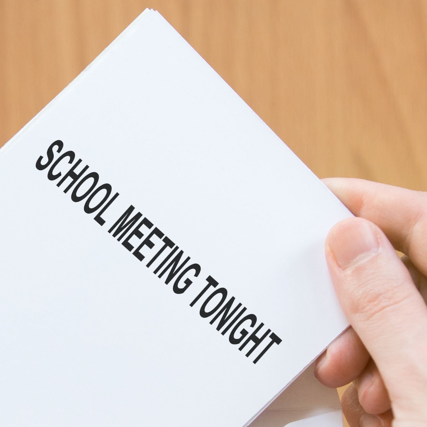 A hand holding a white card stamped with SCHOOL MEETING TONIGHT in bold black letters, using a rubber stamp.