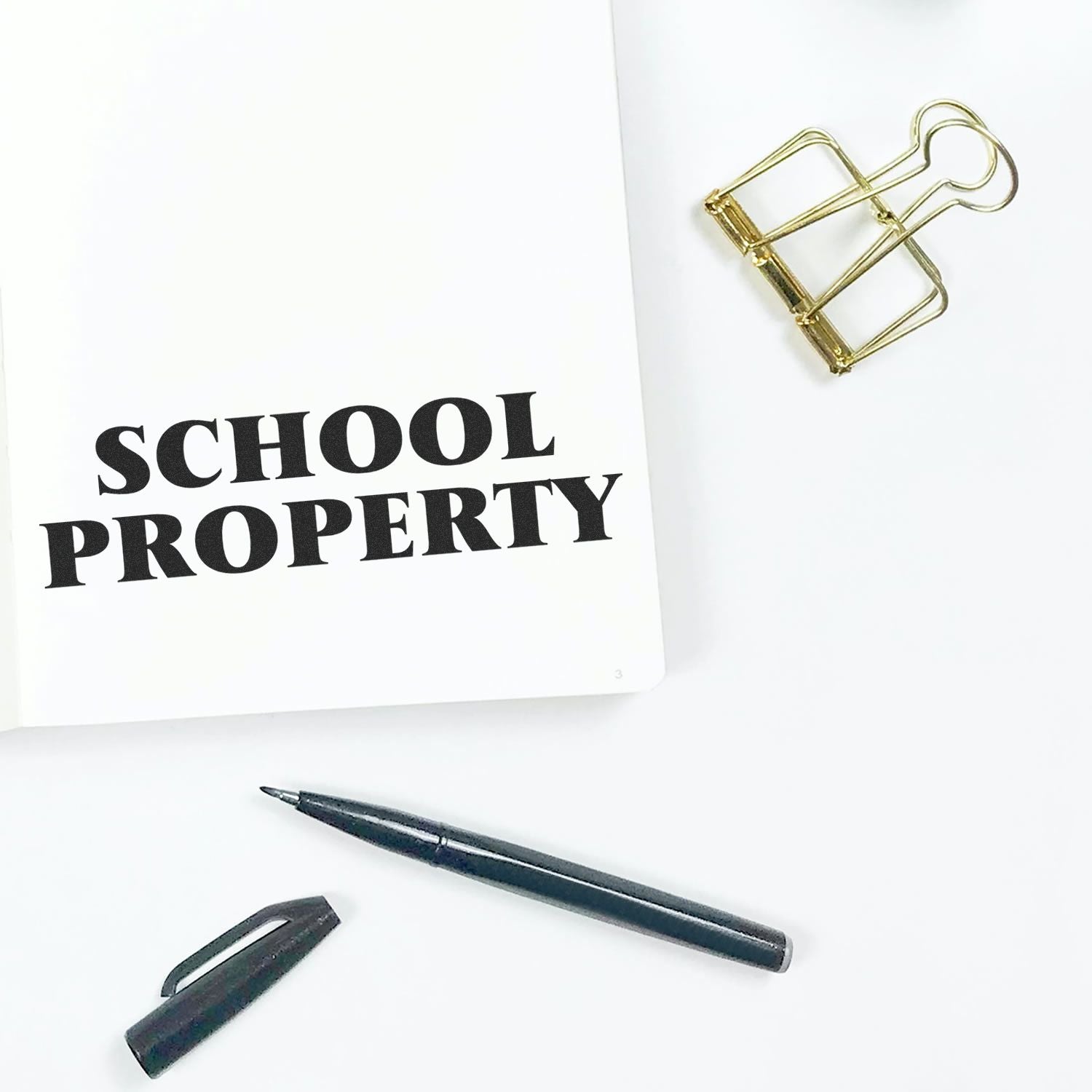 School Property rubber stamp on a white paper with a black pen, pen cap, and a gold binder clip on a white background.