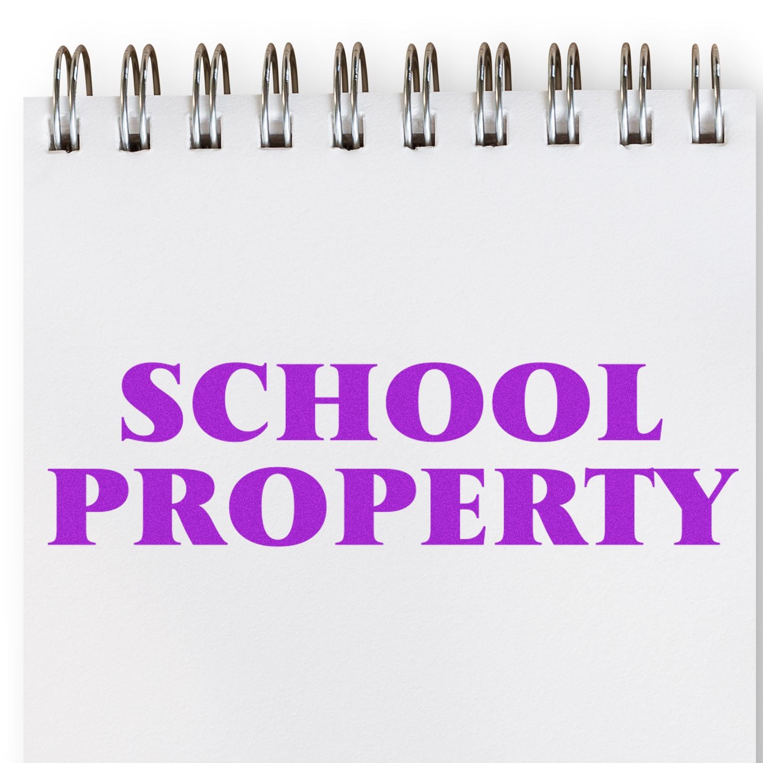 School Property Rubber Stamp in use, imprinting bold purple 'SCHOOL PROPERTY' text on a white spiral-bound notebook page.