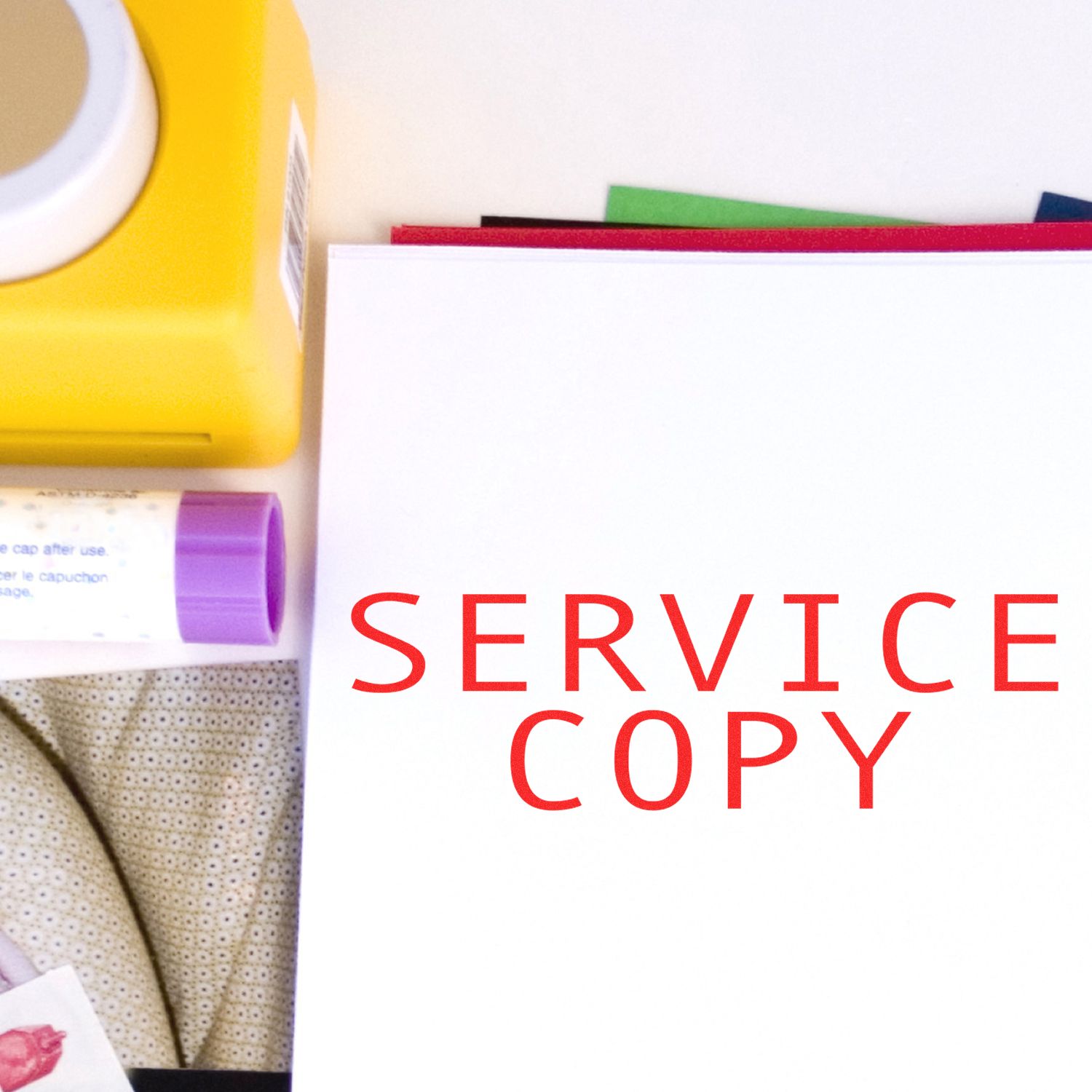 Service Copy rubber stamp in red ink on a white sheet, surrounded by office supplies including a yellow box and glue stick.