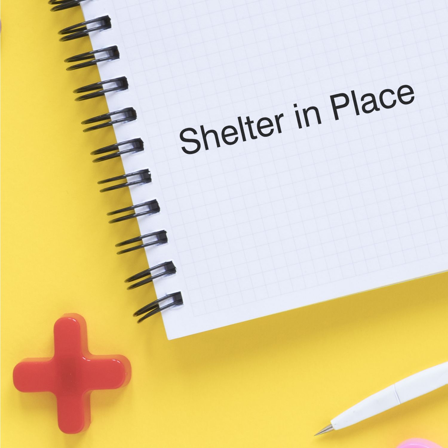 Slim Pre-Inked Shelter in Place Stamp used on a spiral notebook with a red cross and a white pen on a yellow background.