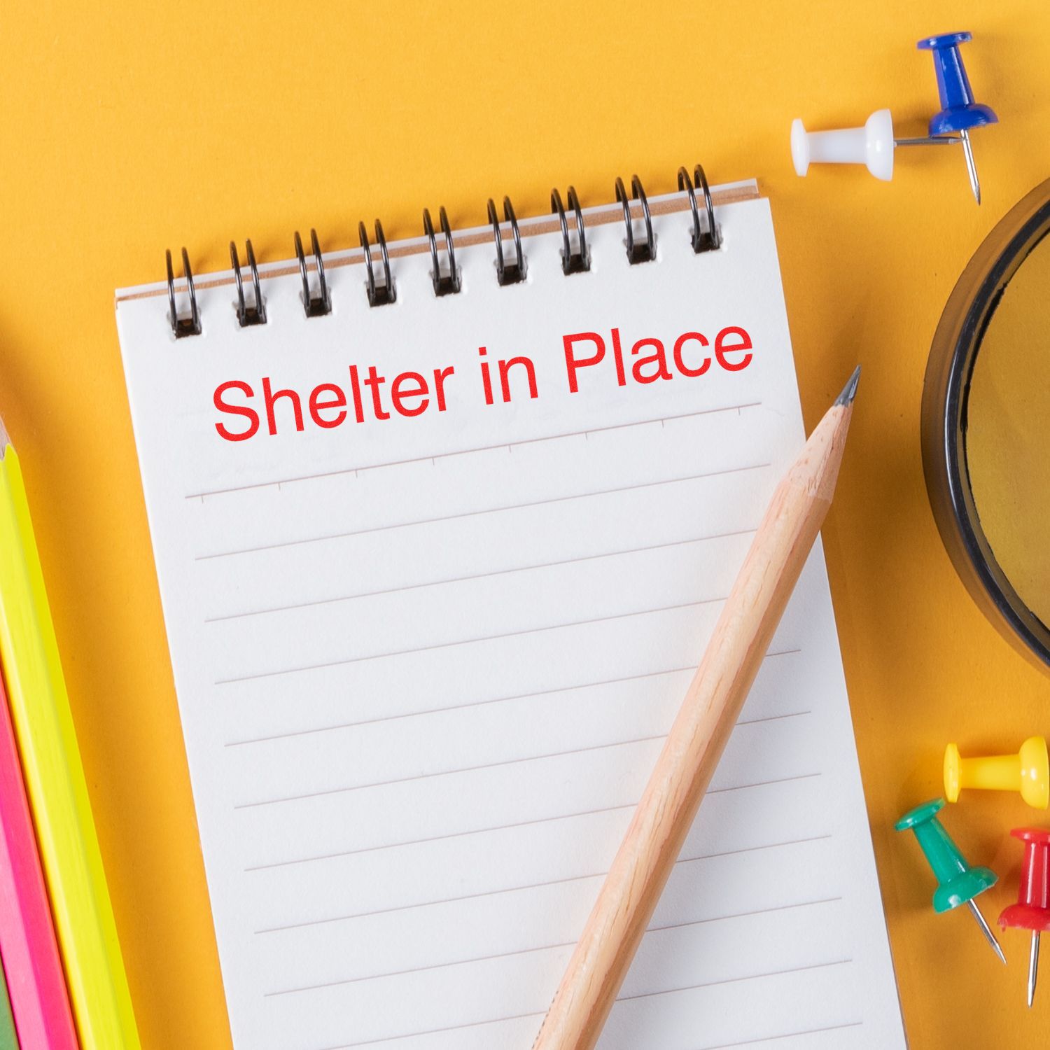 A notepad with Shelter in Place stamped in red, surrounded by colorful push pins, a pencil, and a magnifying glass on a yellow background.