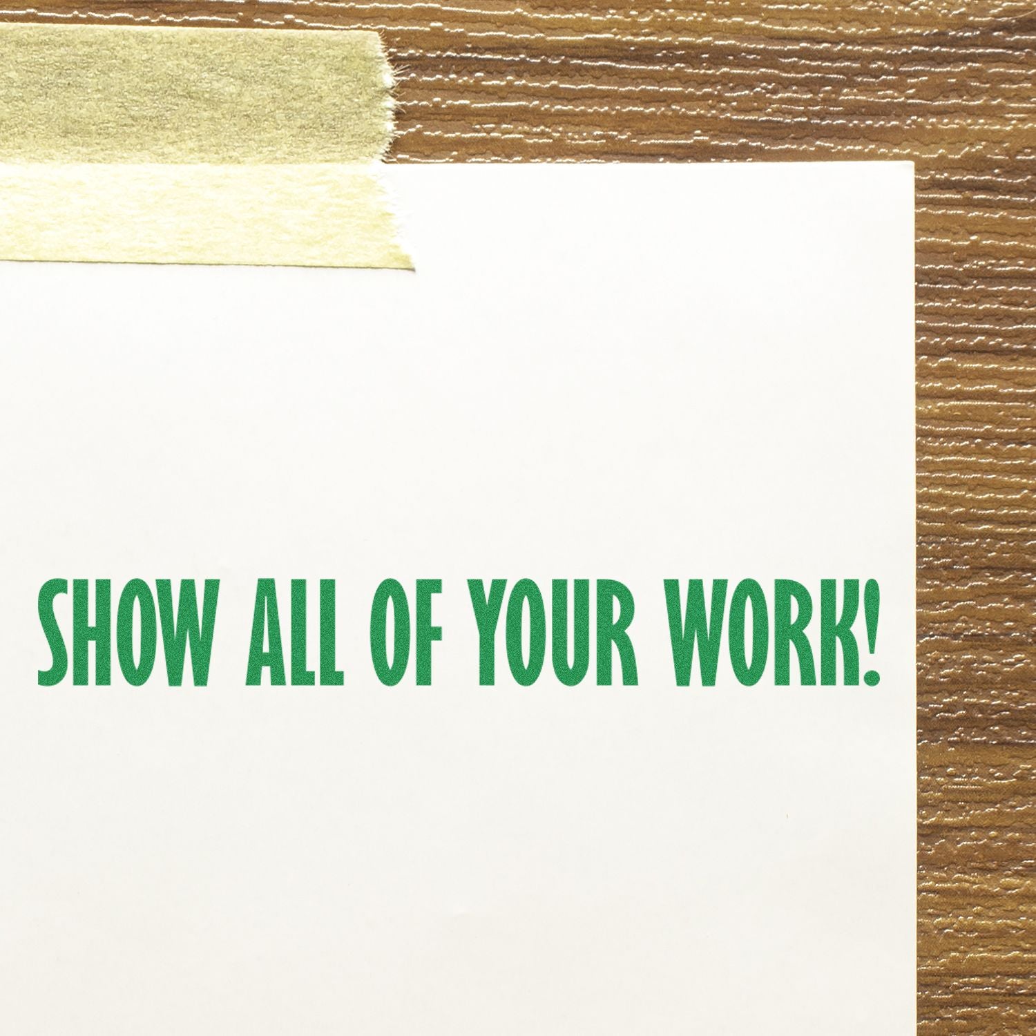 A rubber stamp imprint reading 'Show All Of Your Work!' in green ink on a white paper taped to a wooden surface.