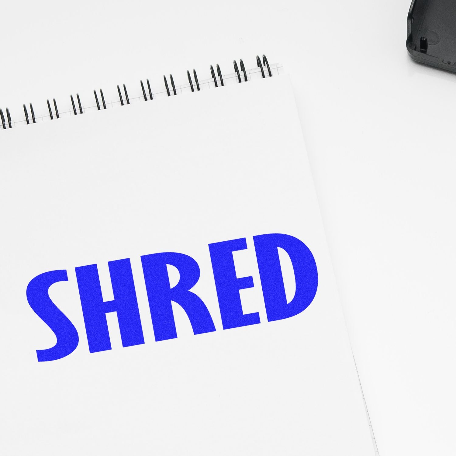 Slim Pre-Inked Shred Stamp imprinting the word 'SHRED' in bold blue letters on a white notepad with a spiral binding.