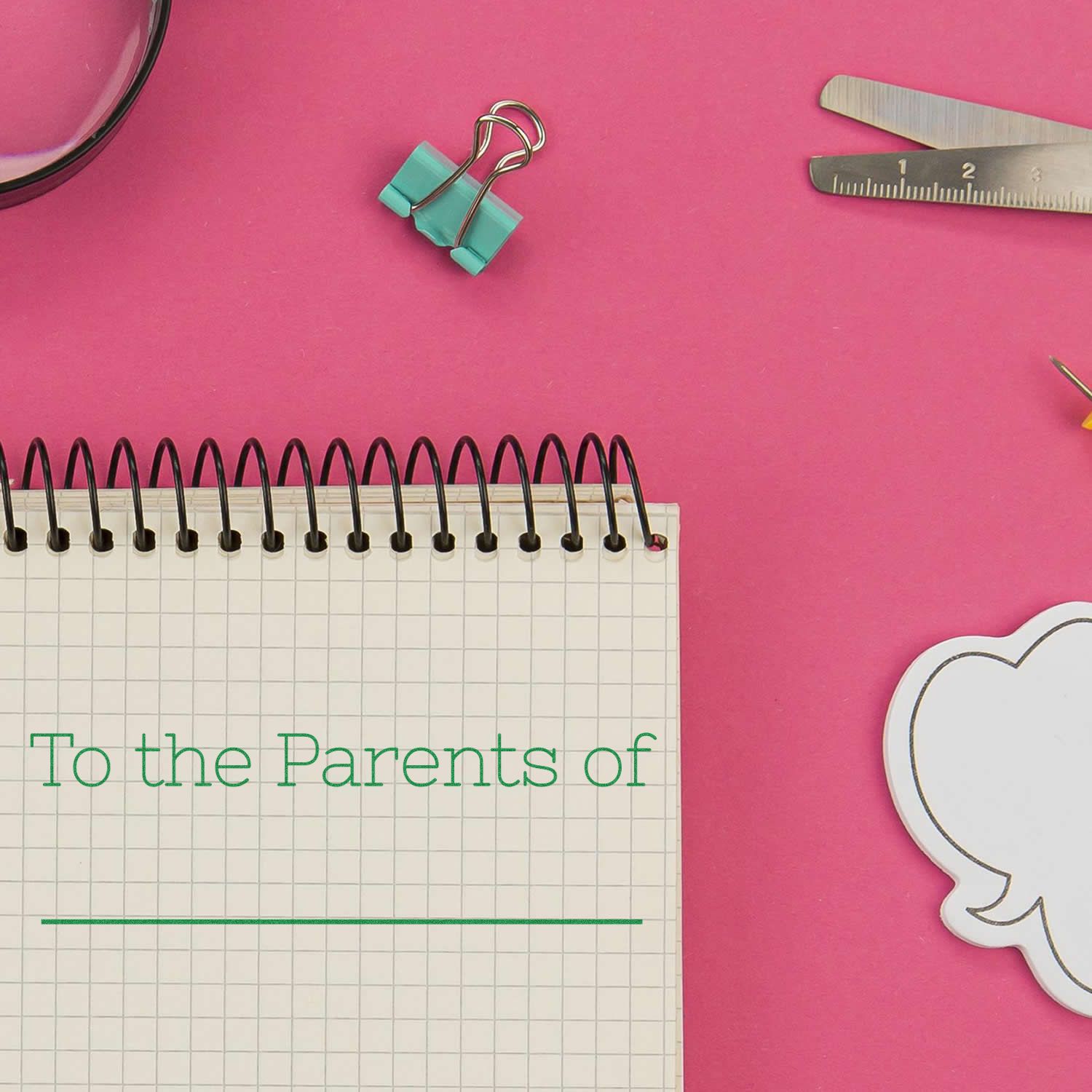 Large Self Inking Skinny To the Parents of Stamp in use on a notebook, surrounded by stationery items on a pink background.