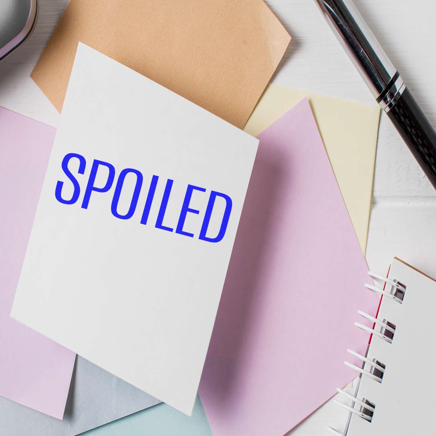 Large Self Inking Spoiled Stamp marking a white paper, surrounded by pastel-colored papers, a pen, and a notebook.