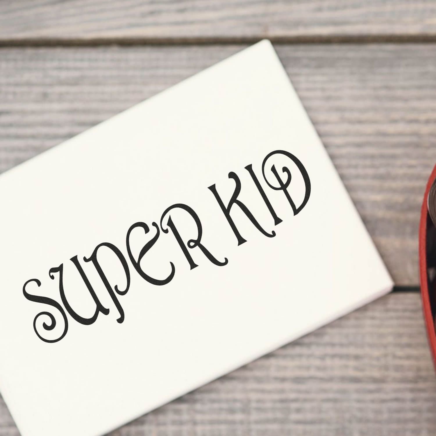 A white paper stamped with SUPER KID using the Self Inking Super Kid Stamp, placed on a wooden surface with part of a red object visible.