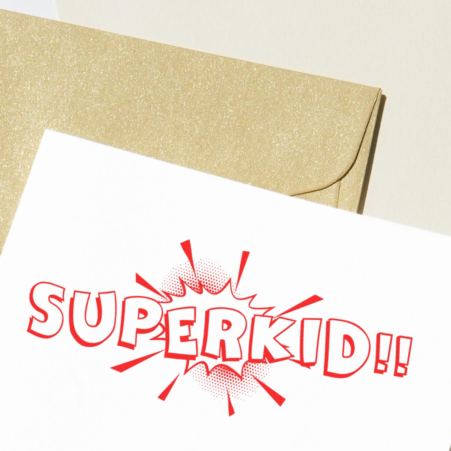 Superkid Rubber Stamp in red ink on white paper with a gold envelope in the background.