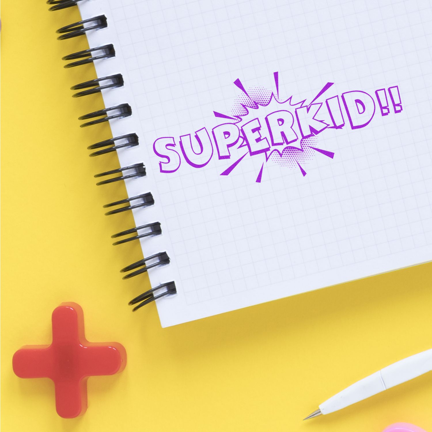 Superkid Rubber Stamp imprint on a spiral notebook with a yellow background, red cross, and white pen nearby.