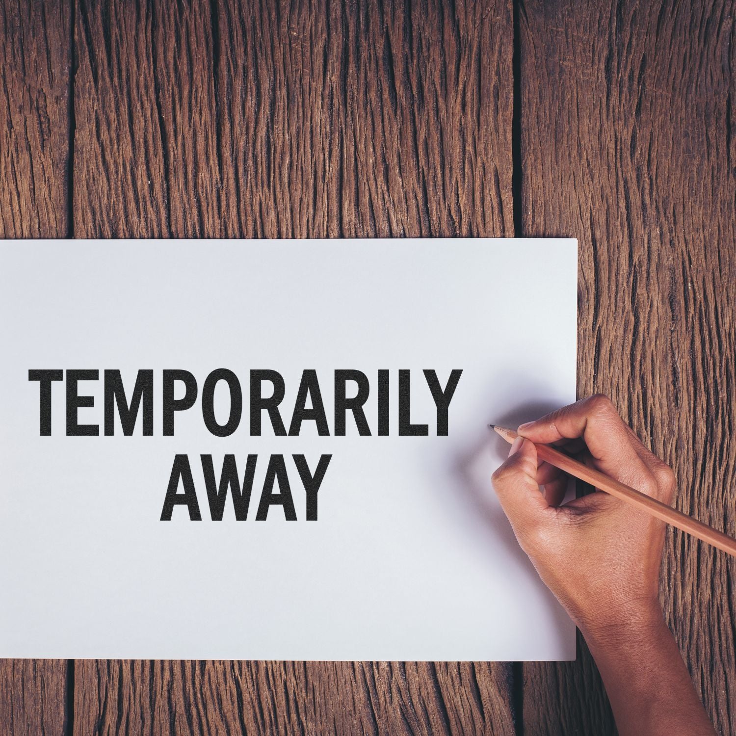 Hand holding a pencil next to a Temporarily Away rubber stamp on a white paper against a wooden background.