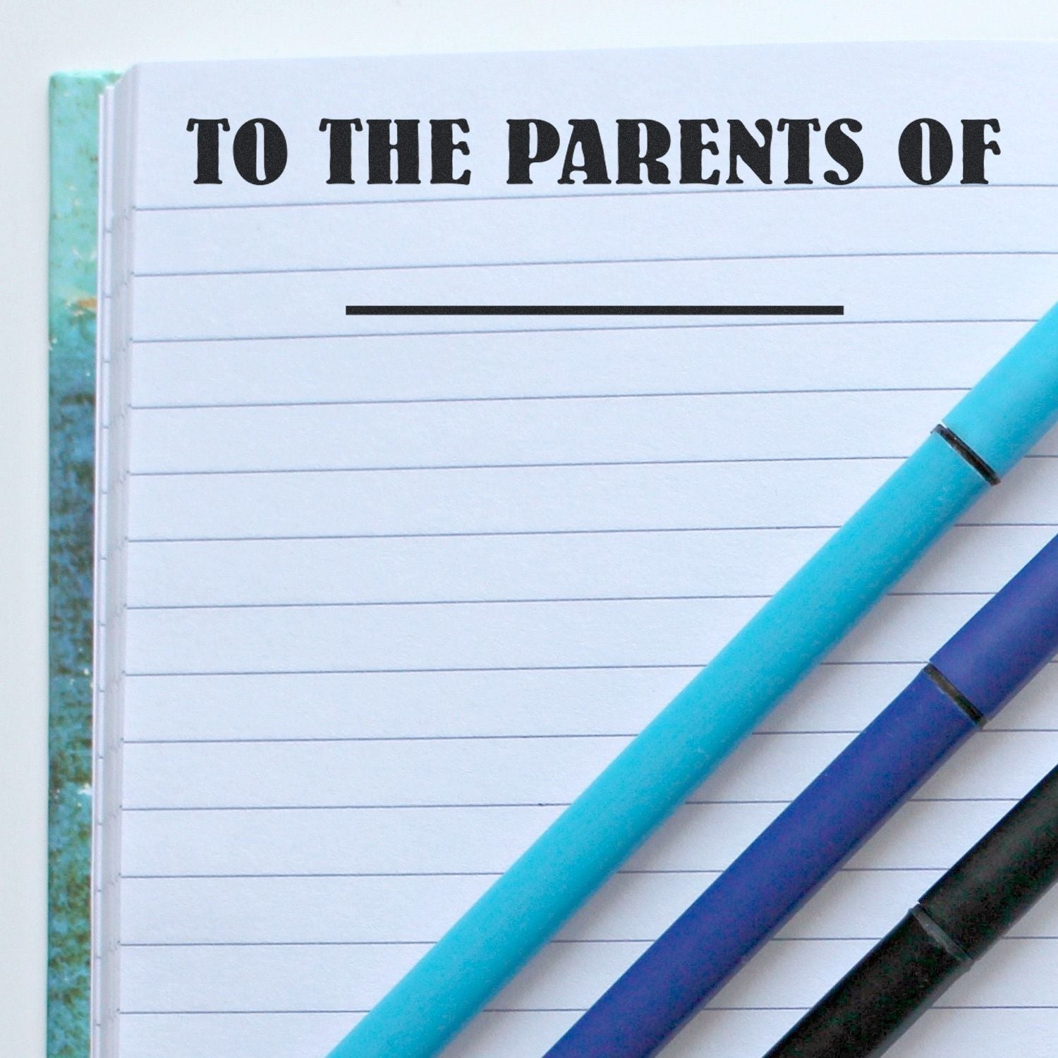 Notebook stamped with TO THE PARENTS OF using the Large Self Inking To The Parents Of Stamp, with three pens placed on top.