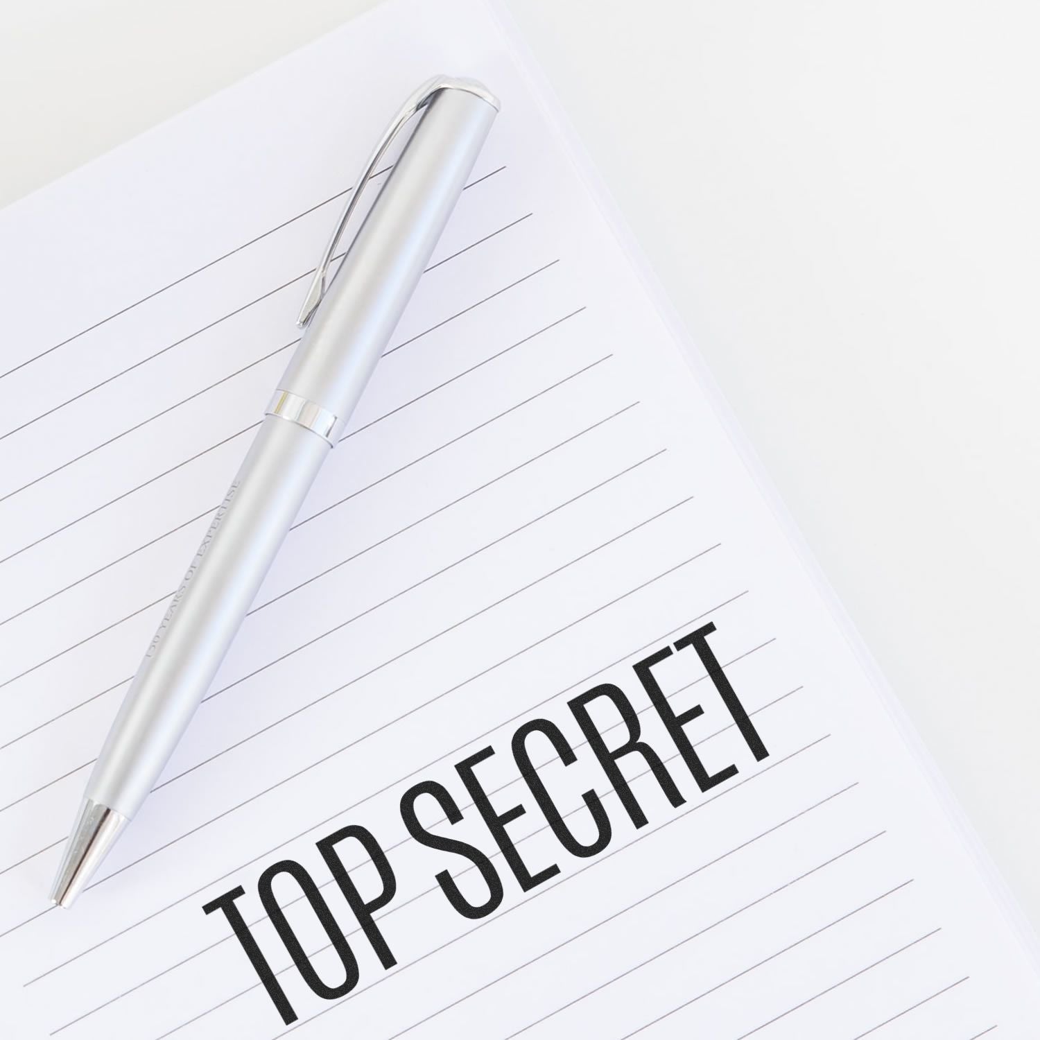 A silver pen rests on a lined notebook with "TOP SECRET" stamped in bold black letters using the Slim Pre-Inked Top Secret Stamp.