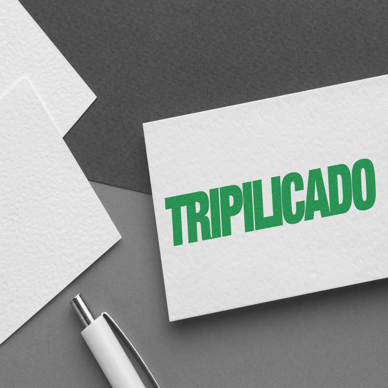 A white card stamped with TRIPILICADO in green ink, placed on a gray surface next to a pen and textured paper.