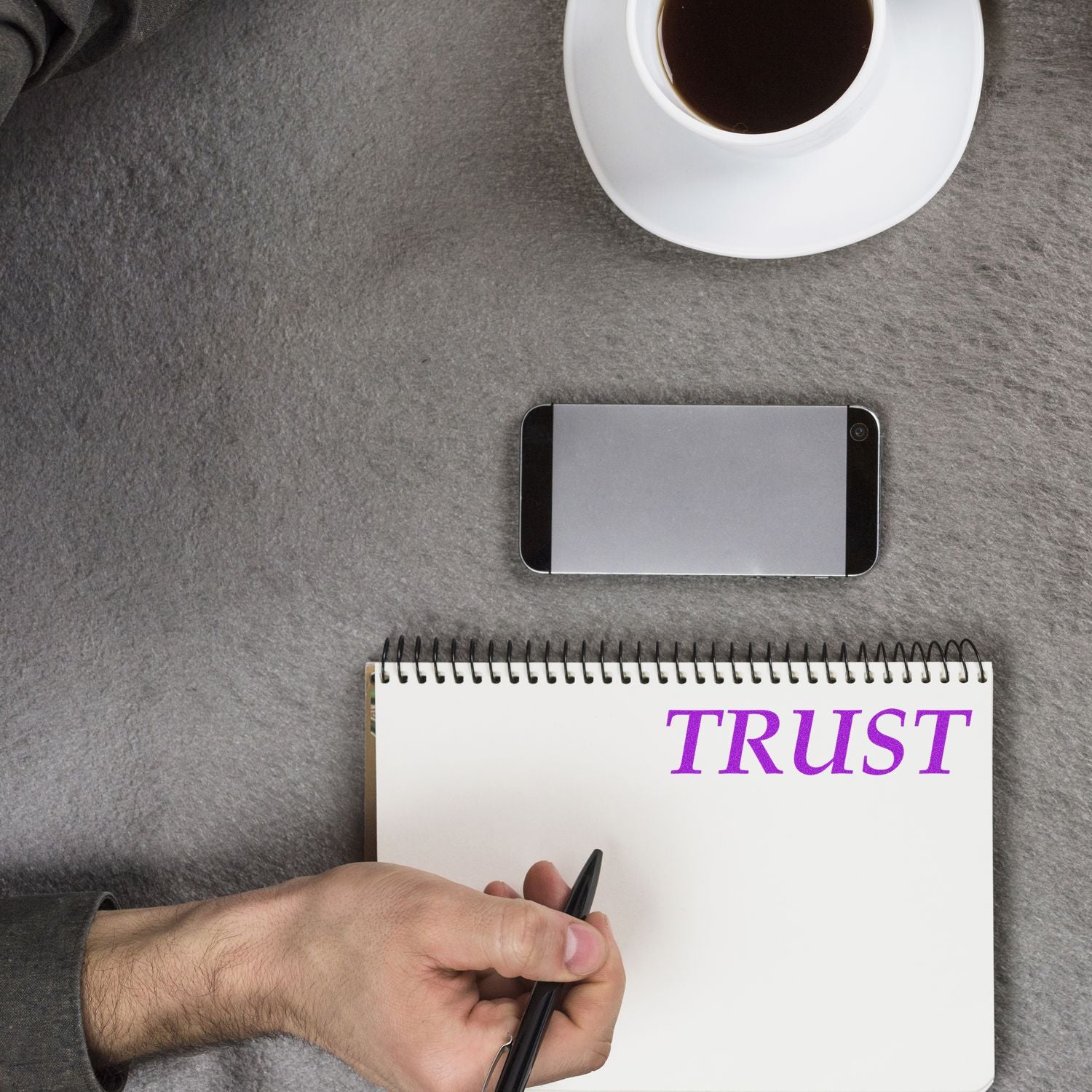 A person using a Self Inking Trust Stamp on a notebook, with a smartphone and a cup of coffee on a gray surface.