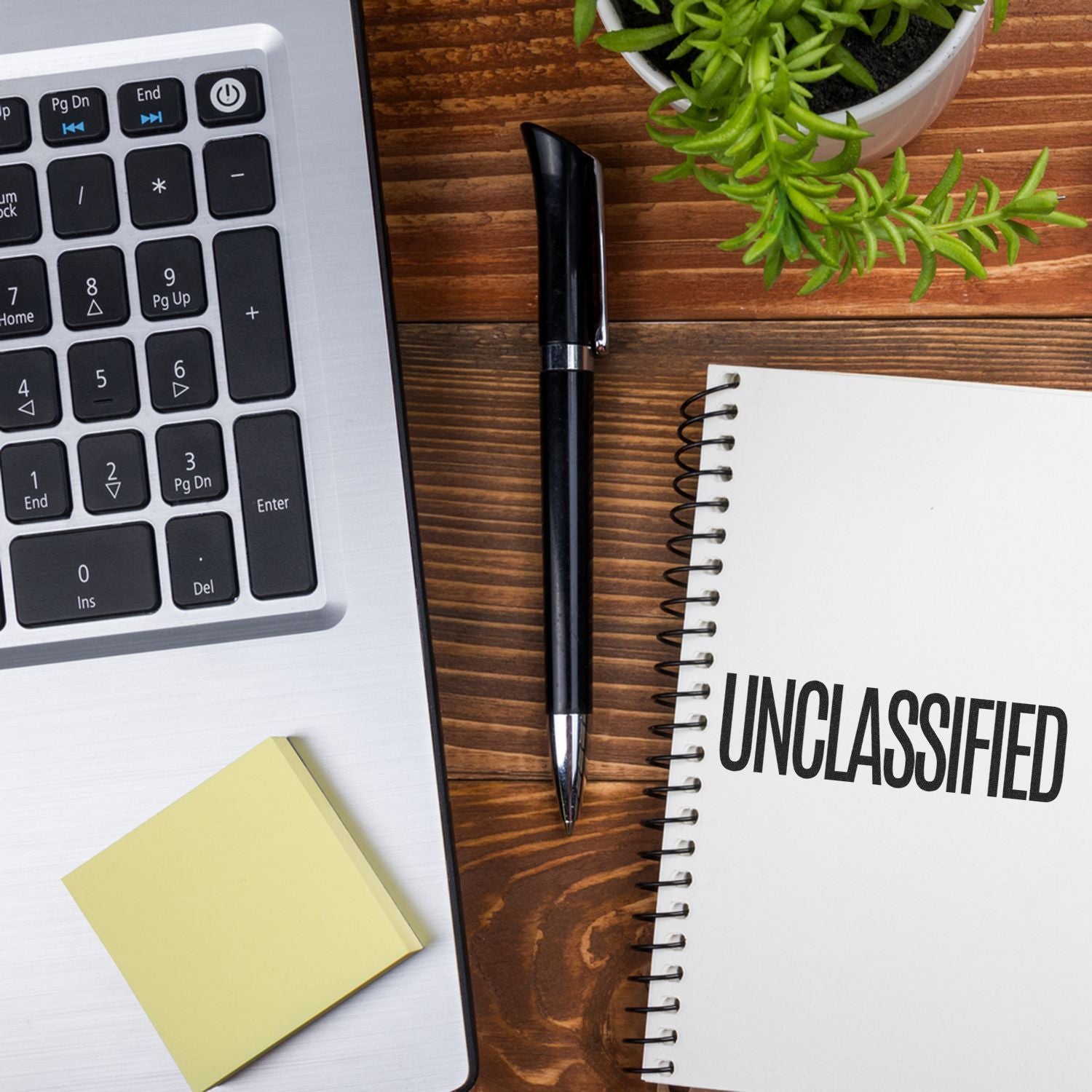 A desk with a laptop, sticky notes, pen, plant, and a notebook stamped with a large unclassified rubber stamp.