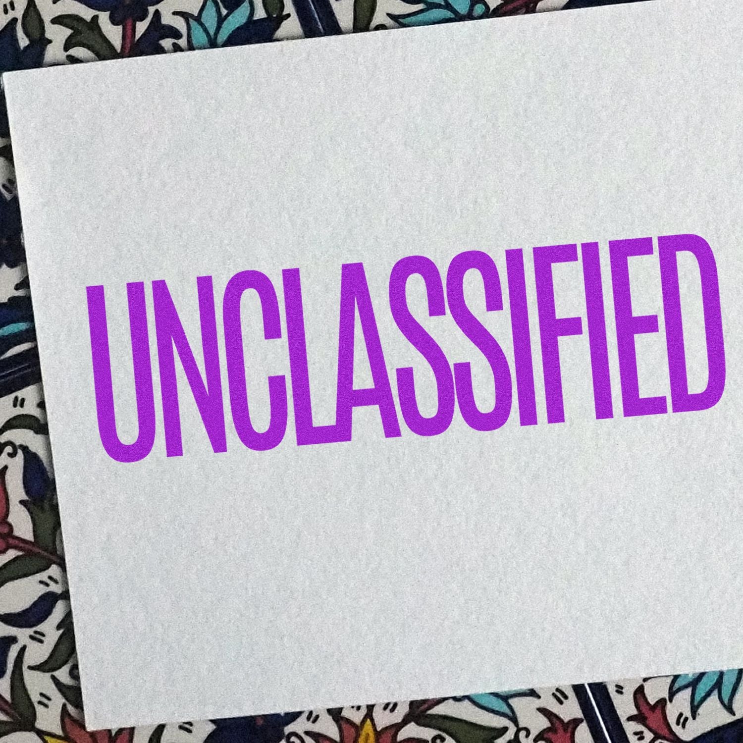 A white paper stamped with UNCLASSIFIED in purple ink using the Self Inking Unclassified Stamp, placed on a floral background.