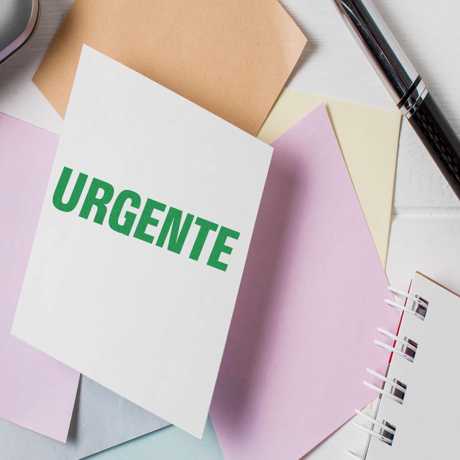 Urgente rubber stamp on white paper surrounded by pastel-colored sheets, a pen, and a notebook on a white desk.