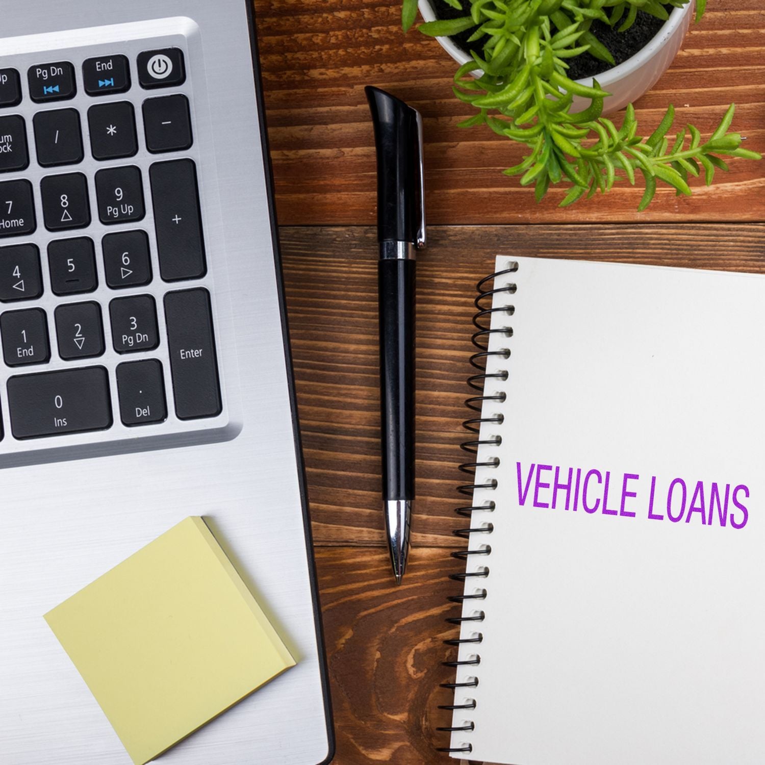 Self Inking Vehicle Loans Stamp next to a laptop, pen, sticky notes, and a notebook with VEHICLE LOANS written on it, placed on a wooden desk.