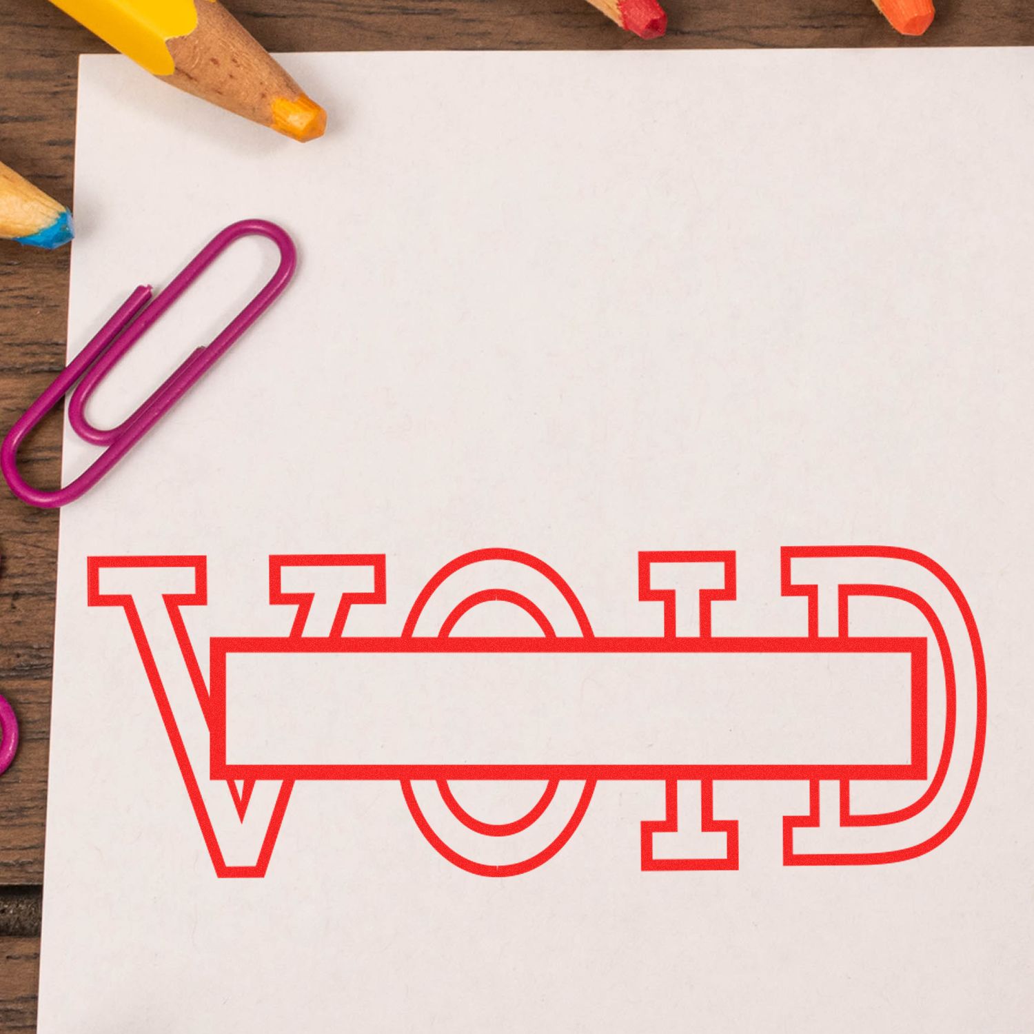 Large Self Inking Void with Box Stamp in red ink on white paper, surrounded by colorful pencils and a purple paperclip on a wooden surface.