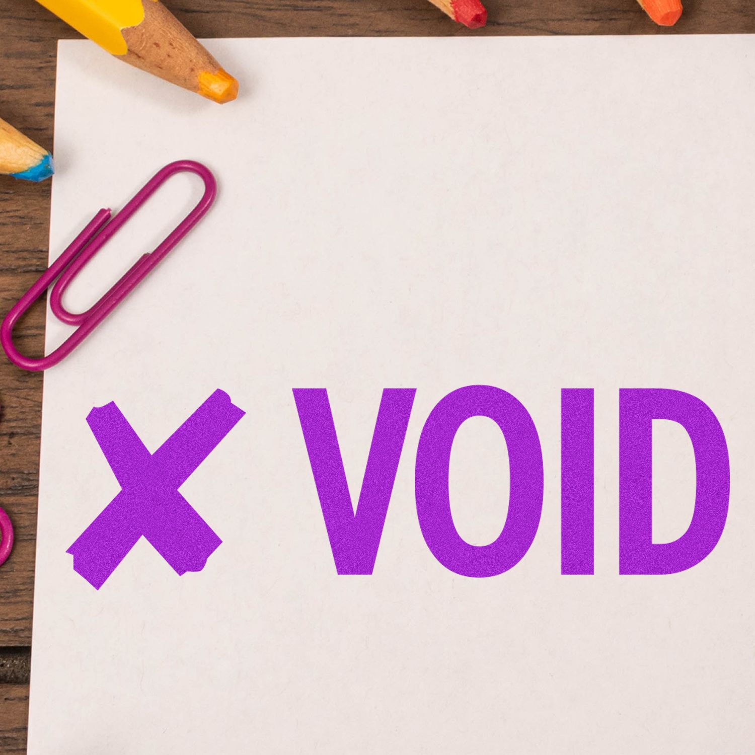 Slim Pre-Inked Void with X Stamp in purple ink on white paper, surrounded by colorful pencils and a paperclip on a wooden surface.