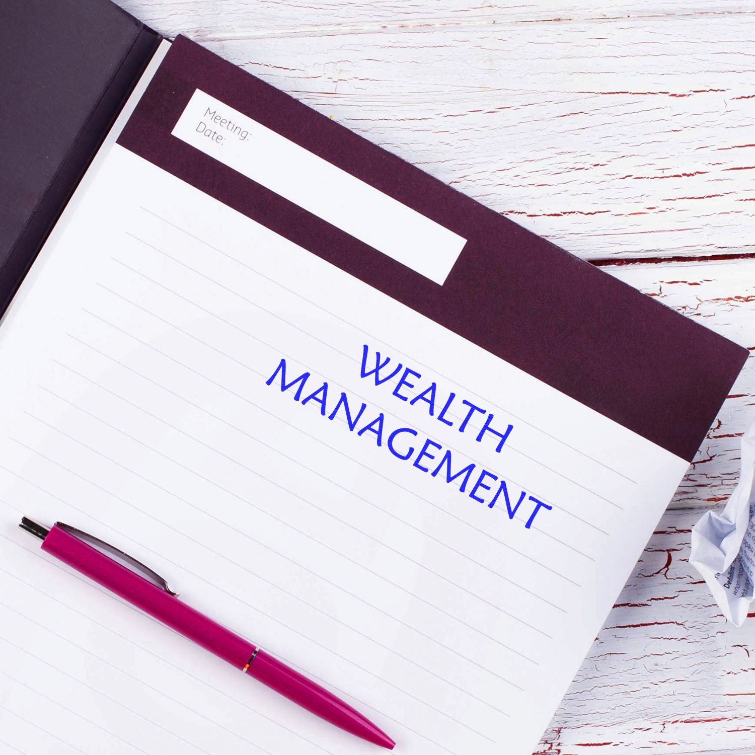 Notebook with Wealth Management stamped in blue ink, a red pen on the left, and a crumpled paper on the right on a white wooden table.