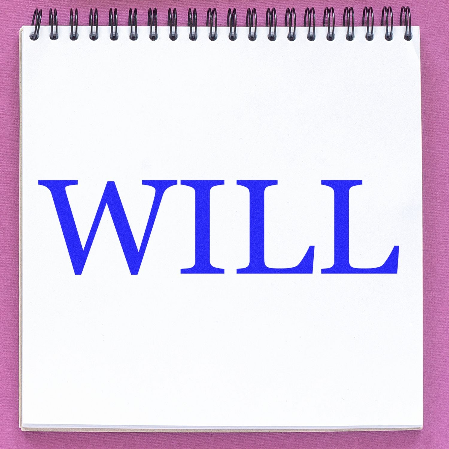 A Self Inking Will Stamp imprints the word WILL in bold blue letters on a white spiral-bound notebook, placed on a pink surface.