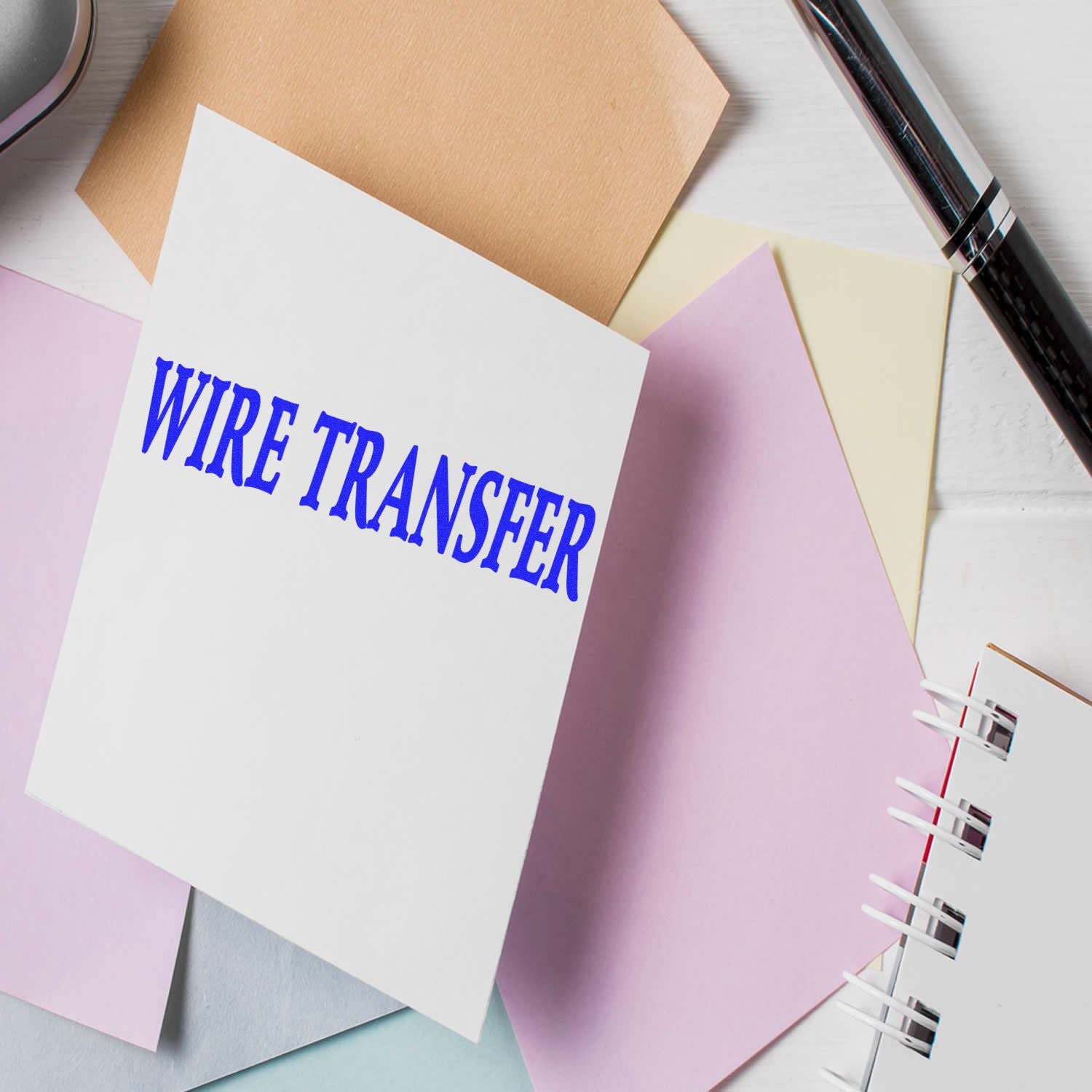 Wire Transfer rubber stamp on a white card surrounded by pastel-colored papers and a pen on a white surface.