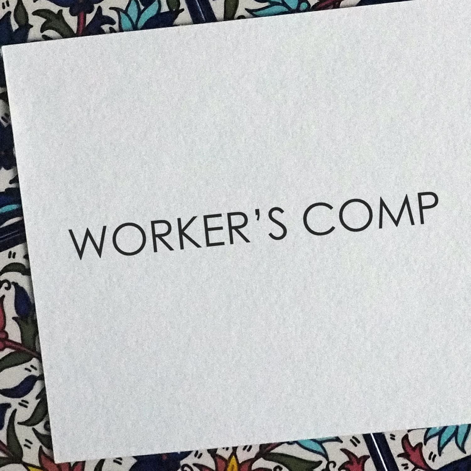 Paper stamped with 'WORKER'S COMP' using a Workers Comp rubber stamp, placed on a colorful patterned surface.