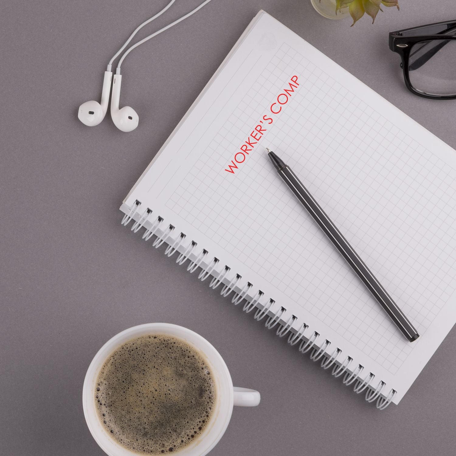 Slim Pre-Inked Workers Comp Stamp on a notebook, surrounded by a pen, coffee cup, glasses, and earphones on a gray desk.