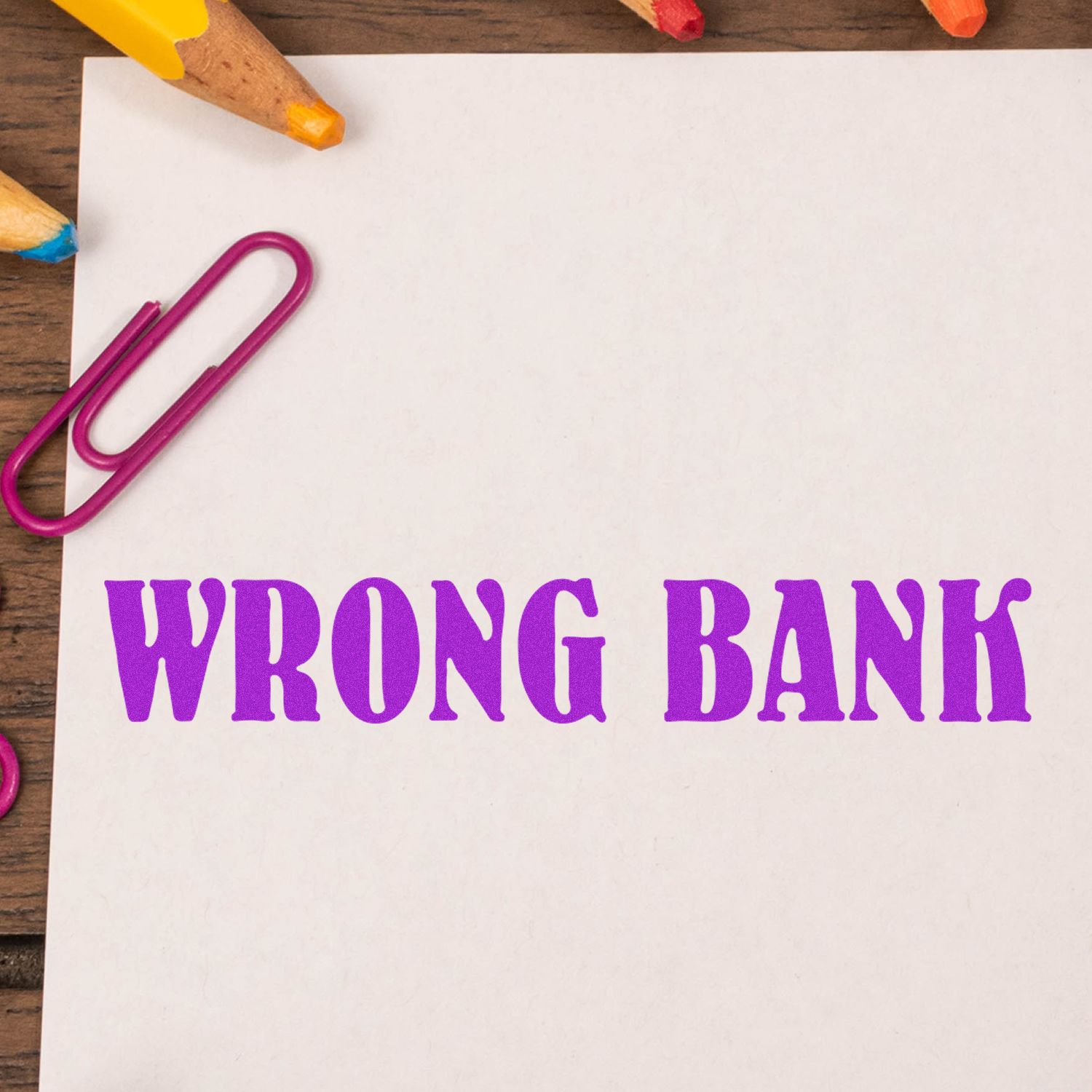 Large Self Inking Wrong Bank Stamp in purple ink on white paper, surrounded by colorful pencils and a pink paperclip on a wooden surface.