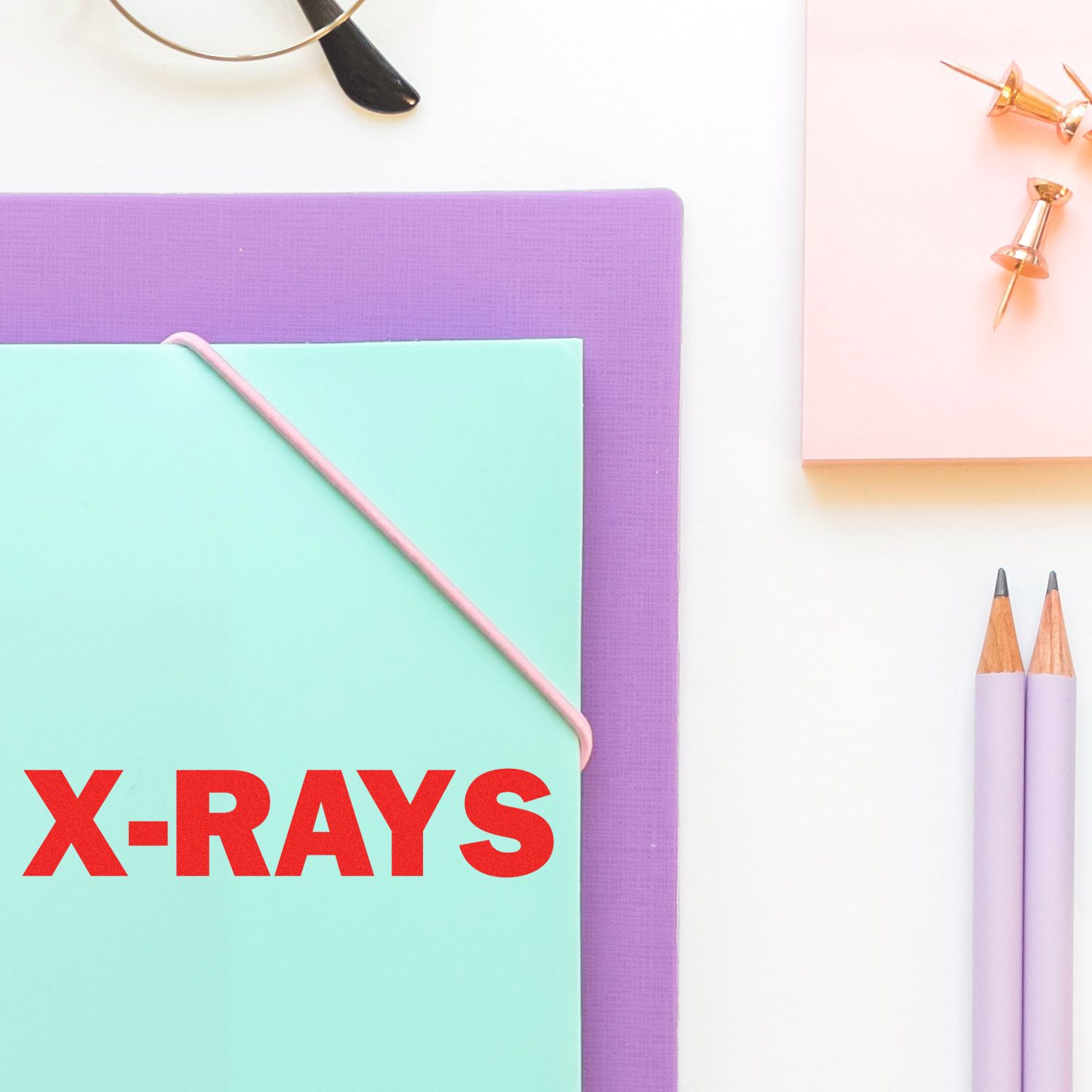 X-Rays rubber stamp on a mint green folder, surrounded by a purple folder, pencils, push pins, and glasses.