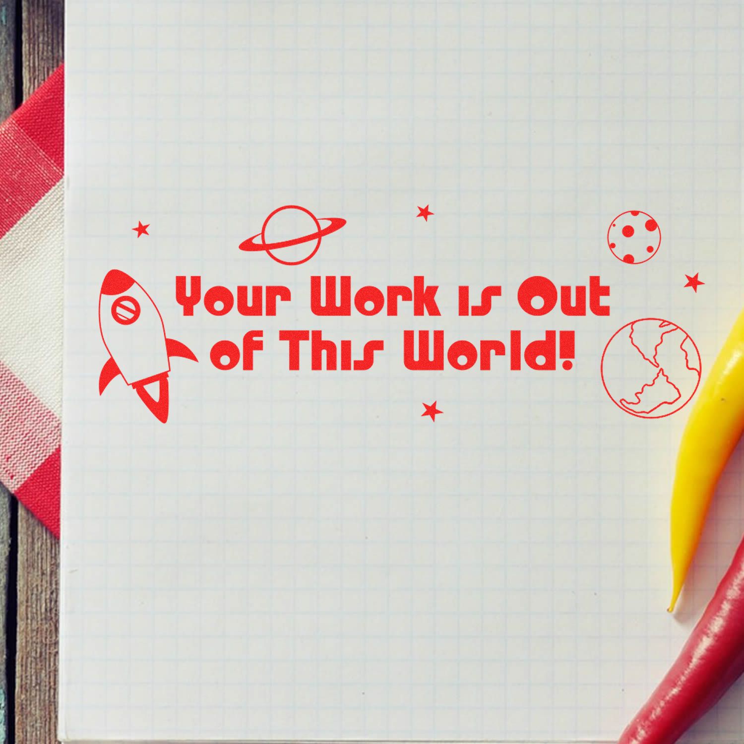 Your Work is Out of This World Rubber Stamp in red ink on graph paper with space-themed icons, next to a yellow chili pepper.