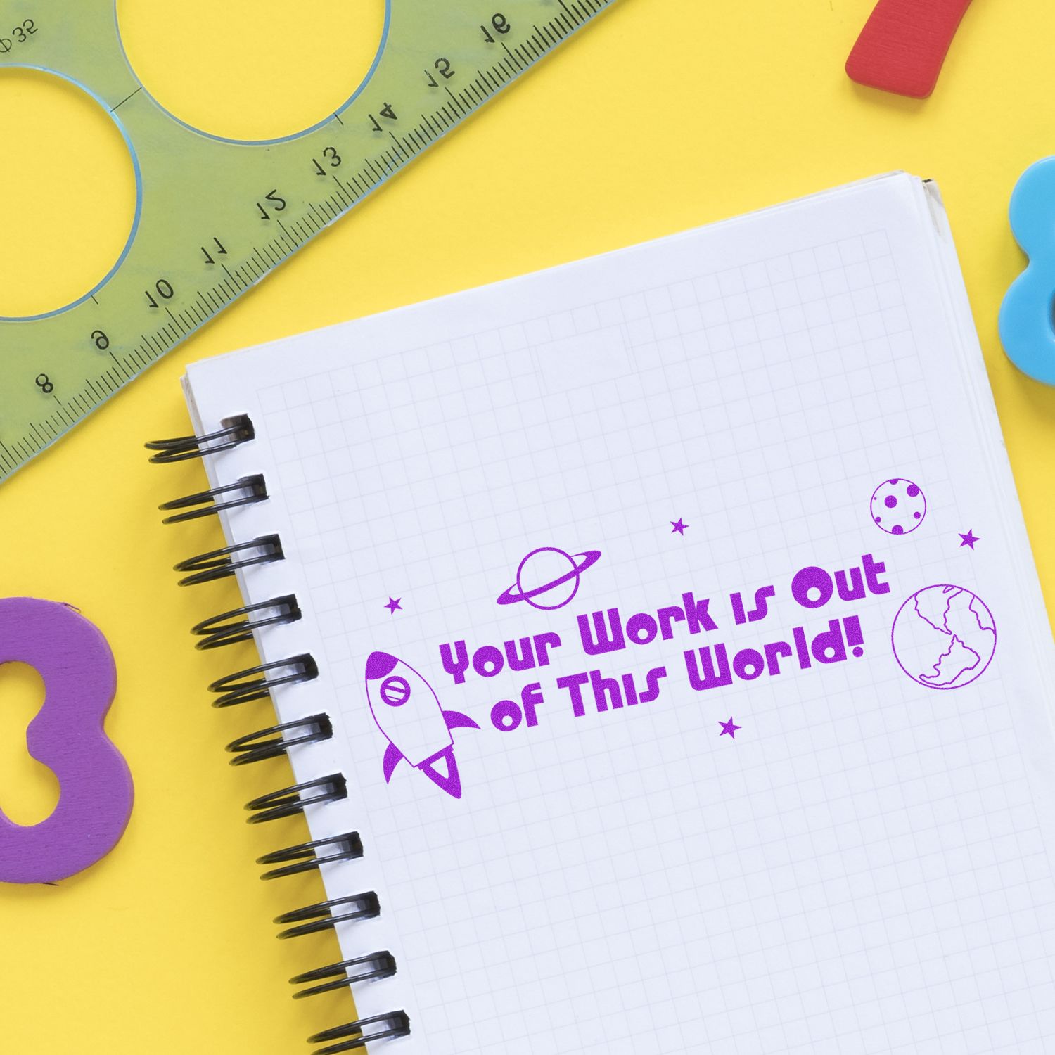 Notebook with a stamped message 'Your Work is Out of This World' in purple ink, surrounded by colorful stationery on a yellow background.