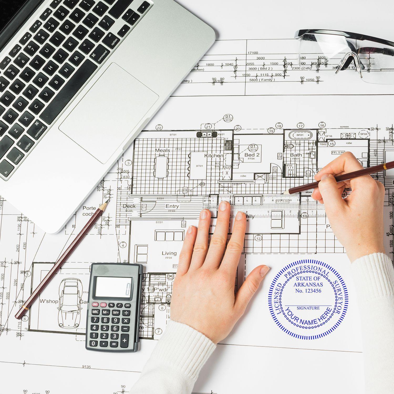 Person using Xstamper Land Surveyor Pre-Inked Rubber Stamp of Seal on architectural blueprint, with a laptop, calculator, and glasses nearby.