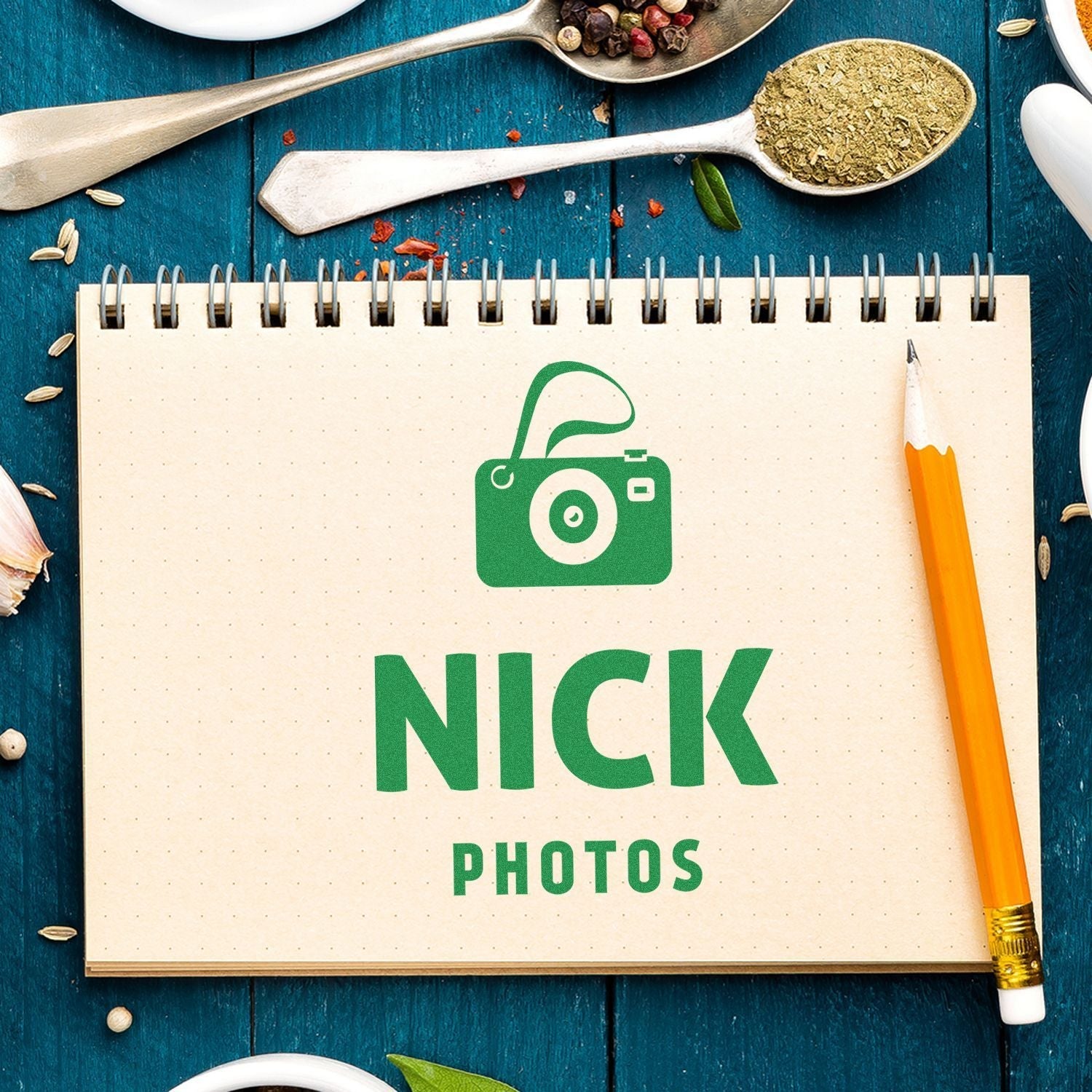 A Custom Rubber Stamp Size 4 x 6 imprinting NICK PHOTOS on a notebook, surrounded by a pencil, spices, and kitchen utensils on a blue surface.