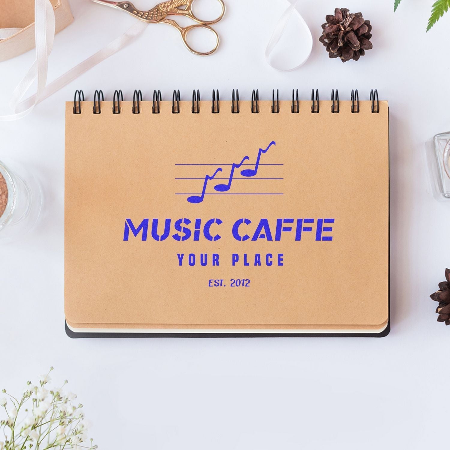 A Custom Rubber Stamp Size 6 x 8 is used to stamp MUSIC CAFFE YOUR PLACE on a spiral notebook, surrounded by stationery items.