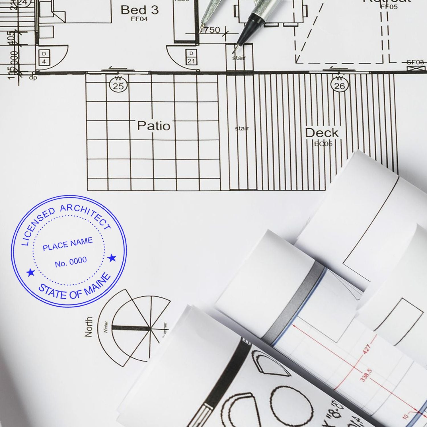 Architectural plans with a Self Inking Maine Architect Stamp, blue ink imprint, and rolled blueprints on a desk.